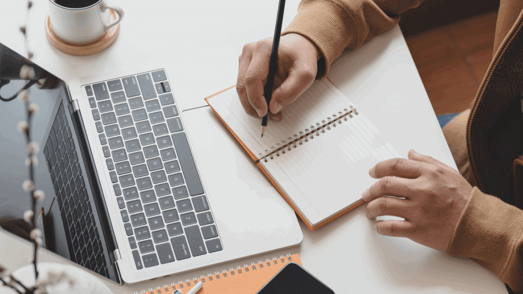Person writing in a notebook next to a laptop