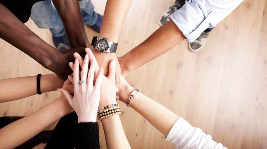 A diverse group of people standing in a circle stack their hands together in the center, symbolizing teamwork, unity, and shared purpose.