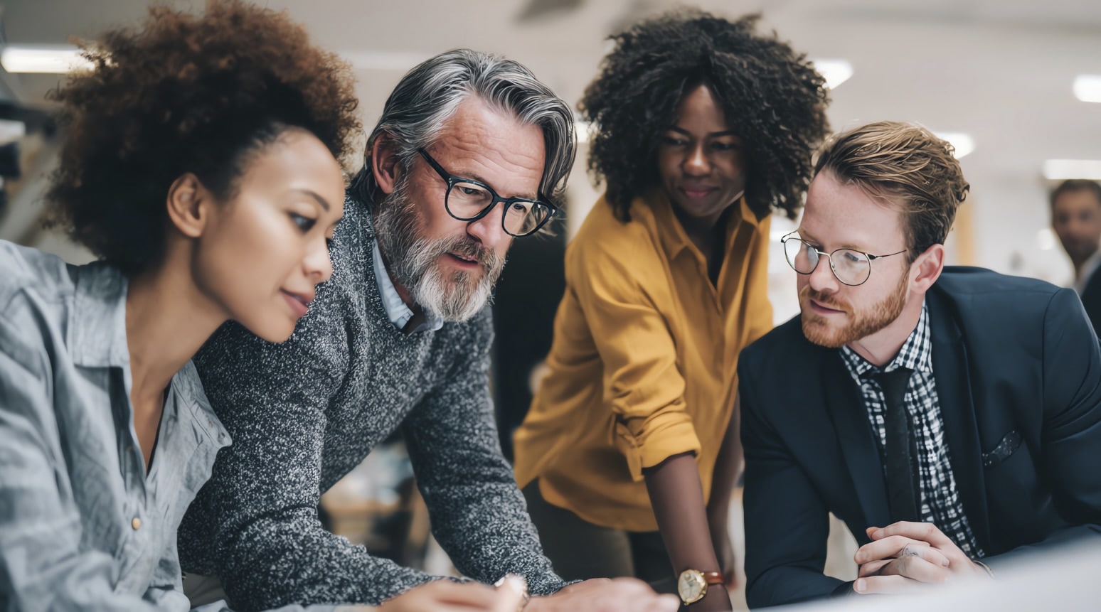 A diverse group of four professionals leaning together over a table, engaged in focused discussion and collaboration.