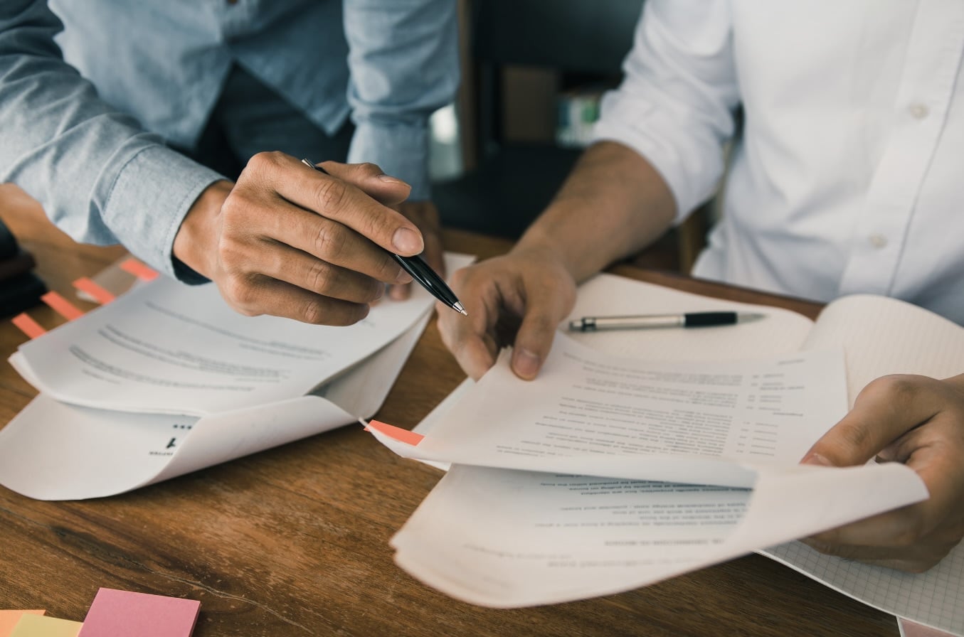 Close-up of two colleagues reviewing printed documents at a wooden desk, one hand pointing a pen at a line of text while sticky note tabs mark other pages for follow-up.