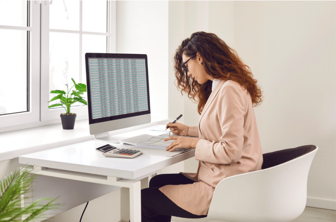 A woman with curly hair in a blazer sits at a white desk reviewing spreadsheet data on a desktop monitor, with a calculator, notepad, and pen in front of her and a potted plant by the window.