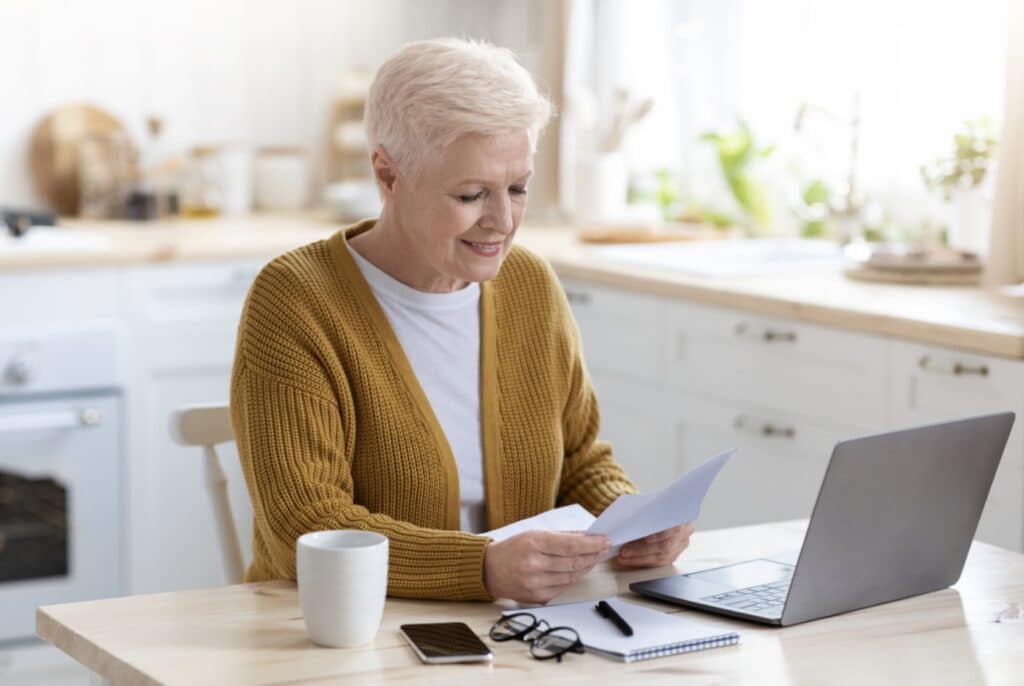 Older woman with short white hair, in a mustard cardigan, smiles while reading a letter at her kitchen table, with a laptop, coffee mug, notebook and glasses beside her.