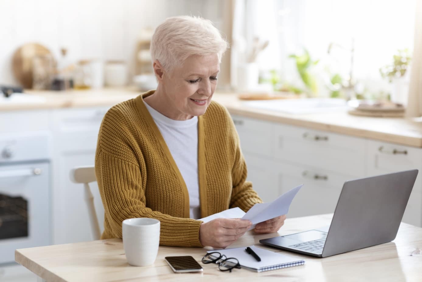 Older woman with short white hair, in a mustard cardigan, smiles while reading a letter at her kitchen table, with a laptop, coffee mug, notebook and glasses beside her.